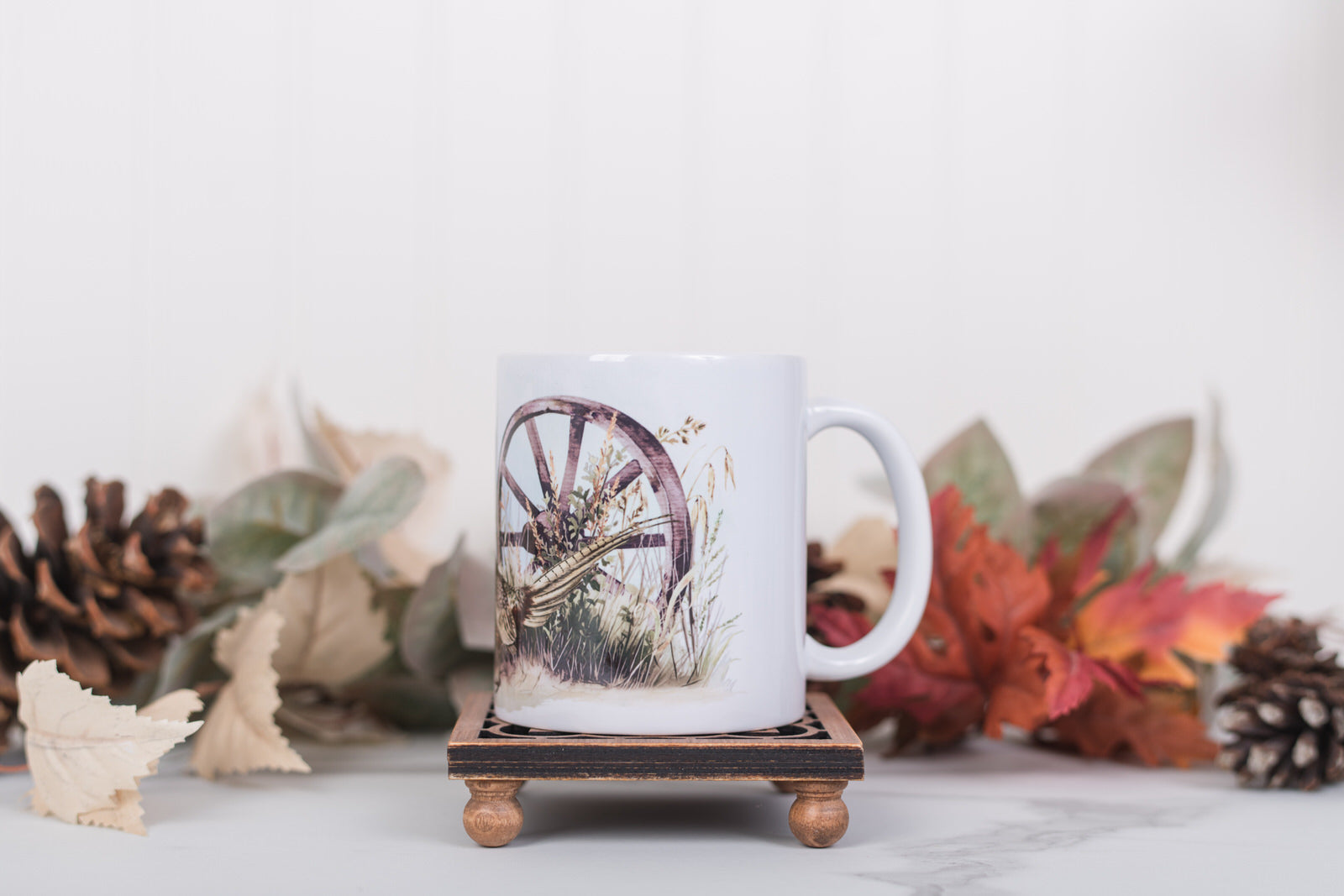 White mug with a wagon wheel design on a wooden coaster with fall decorations in the background.