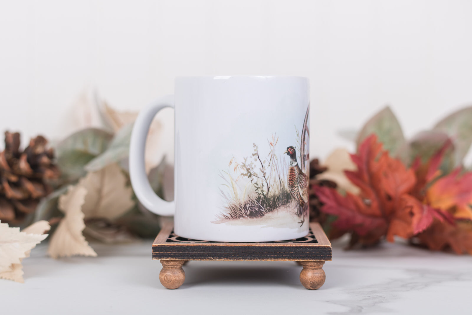 White mug with nature-themed design on a wooden stand with fall leaves and pinecones in the background