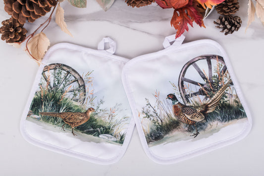 Pair of pot holders with pheasant design on a white surface with autumn decorations.