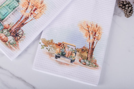 Towels with autumn-themed designs featuring trees, pumpkins, and a truck on a white surface.