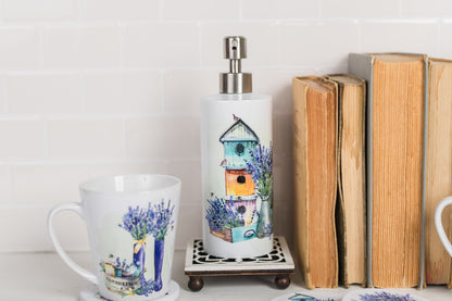 A stainless steel soap dispenser with a French lavender design, featuring colorful watercolour patterns with birdhouses, placed on a wooden stand next to a mug and moldy books.