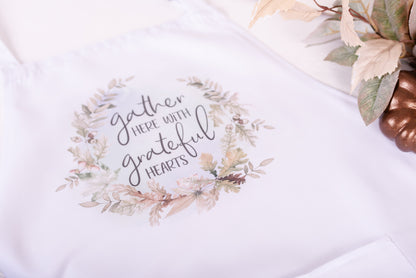 A white apron with a floral design around the neckline and the text 'gather here with grateful hearts' printed on the front bib.