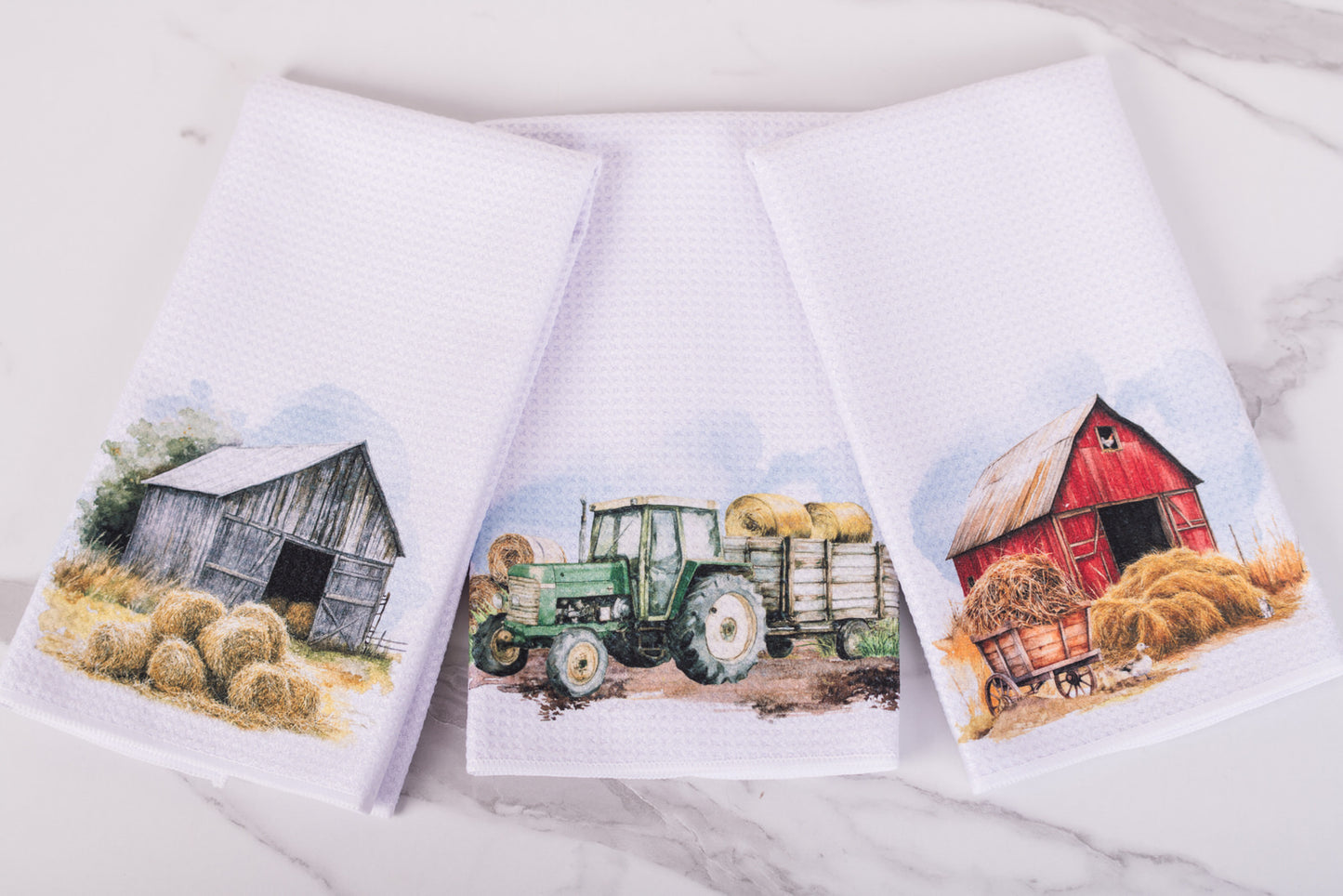 Three towels with farm-themed designs featuring a barn, tractor, and wagon on a marble counter.