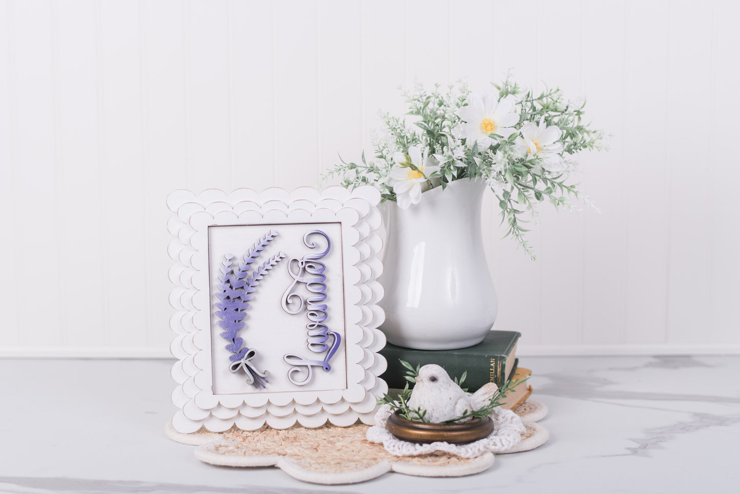 Decorative items including a framed lavender DIY wood piece with floral designs, a white vase with greenery, and a small white bird figurine on a light surface.