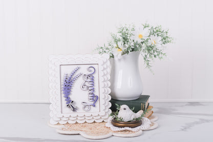 Decorative items including a framed lavender DIY wood piece with floral designs, a white vase with greenery, and a small white bird figurine on a light surface.