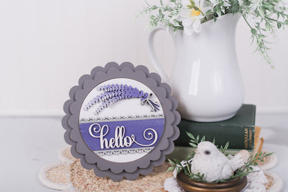 Decorative sign with 'hello' and lavender design on a white surface with a vase and books in the background.