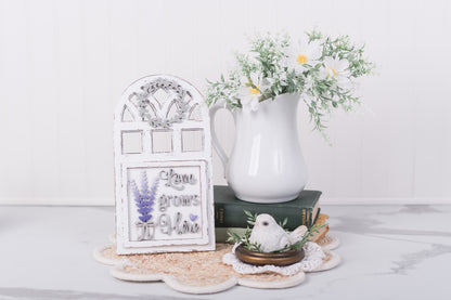 Decorative items including a white pitcher, lavender framed craft, and small bird figurine on a white surface.