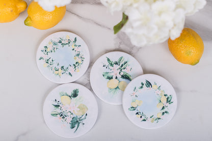 Set of four round coasters with lemon designs on a light surface with lemons and flowers.