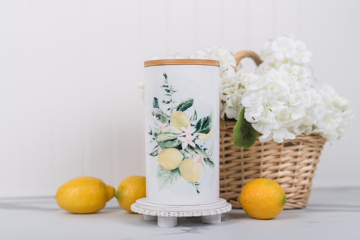 Large ceramic kitchen cannister with lemons. Styled on a marble countertop with a white bead board, backsplash paired with a basket of flowers and lemons.