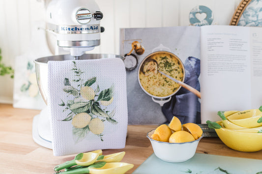 Kitchen scene with a KitchenAid mixer, lemon-themed towel, cookbook, and lemons on a wooden table.