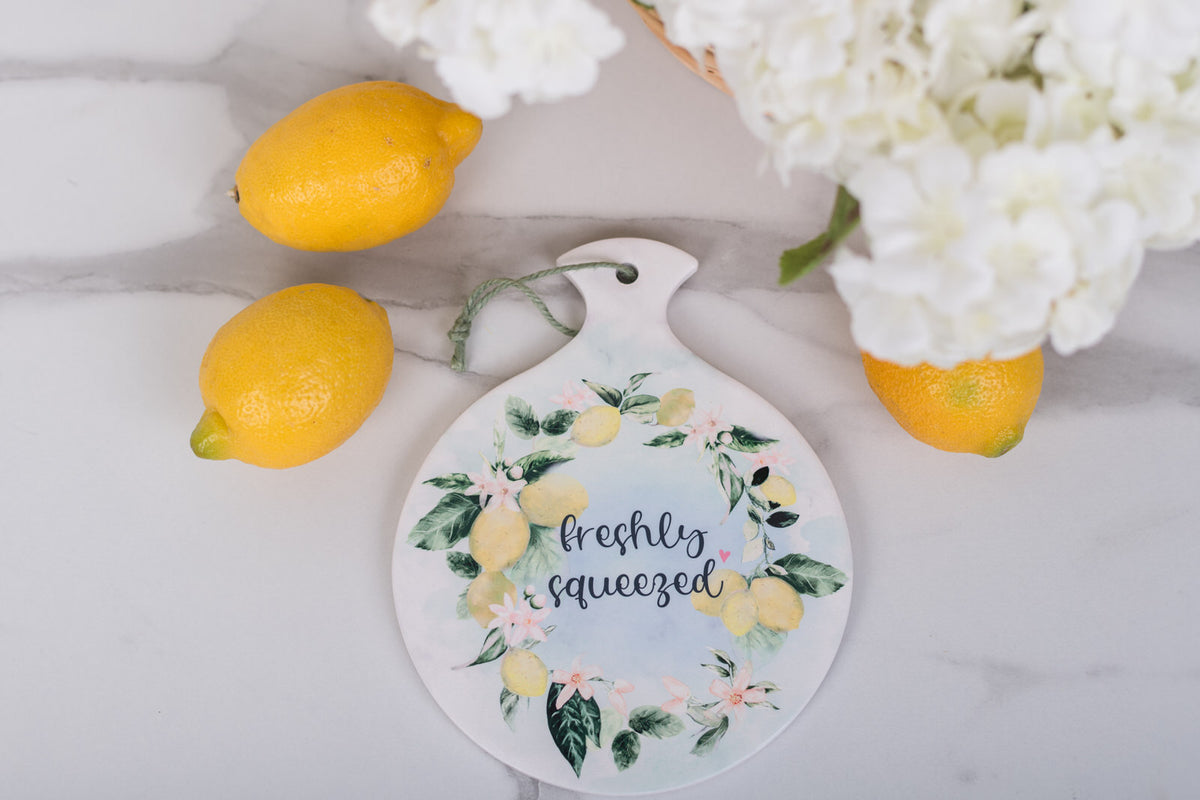 Decorative trivet with lemon design and text, surrounded by lemons and white flowers on a marble surface.