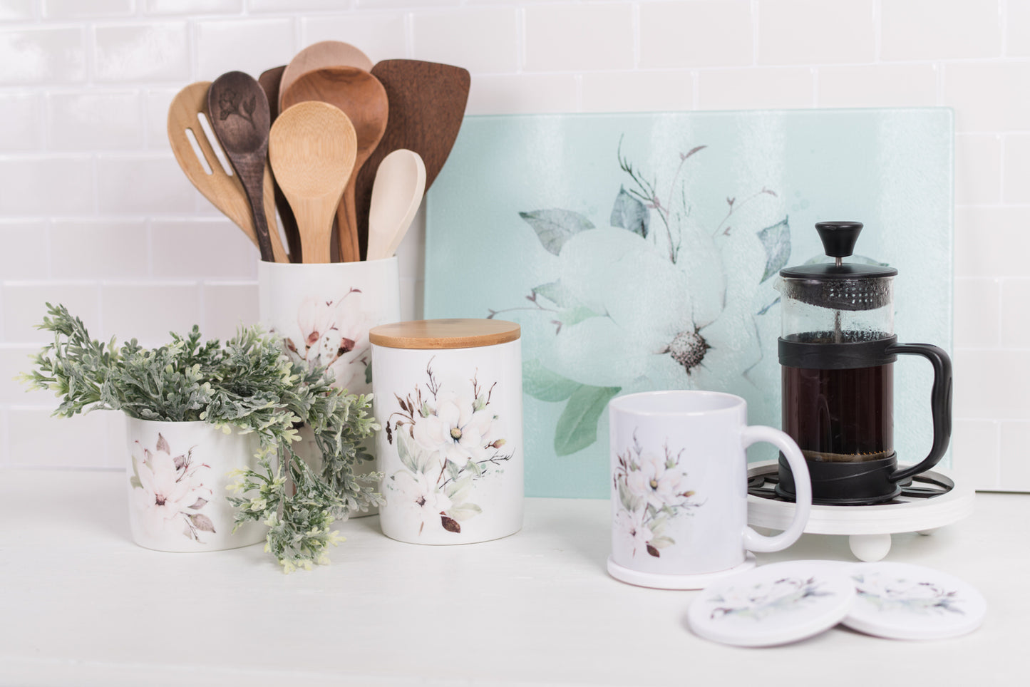 Kitchen scene with floral-patterned mugs, canisters, wooden spoons, and a French press on a white surface.