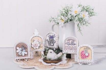 Decorative spring-themed miniature arch signs with flowers and a bird on a white surface.