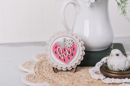 Decorative strawberry sign with 'hi' text on a white surface with a white pitcher and greenery in the background.