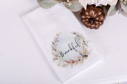 A white hand towel with a green and brown floral pattern and the word 'Thankful' written in the center. The towel is folded and displayed with a fall-colored decor in the background.