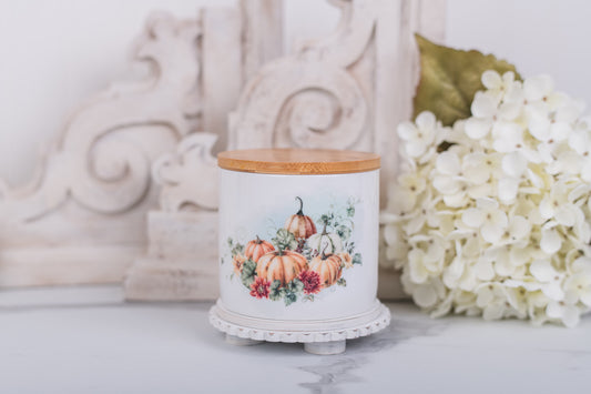Decorative canister with pumpkin design on a white surface with flowers in the background