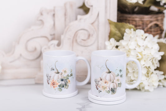 Two white mugs with pumpkin and floral designs on a marble counter