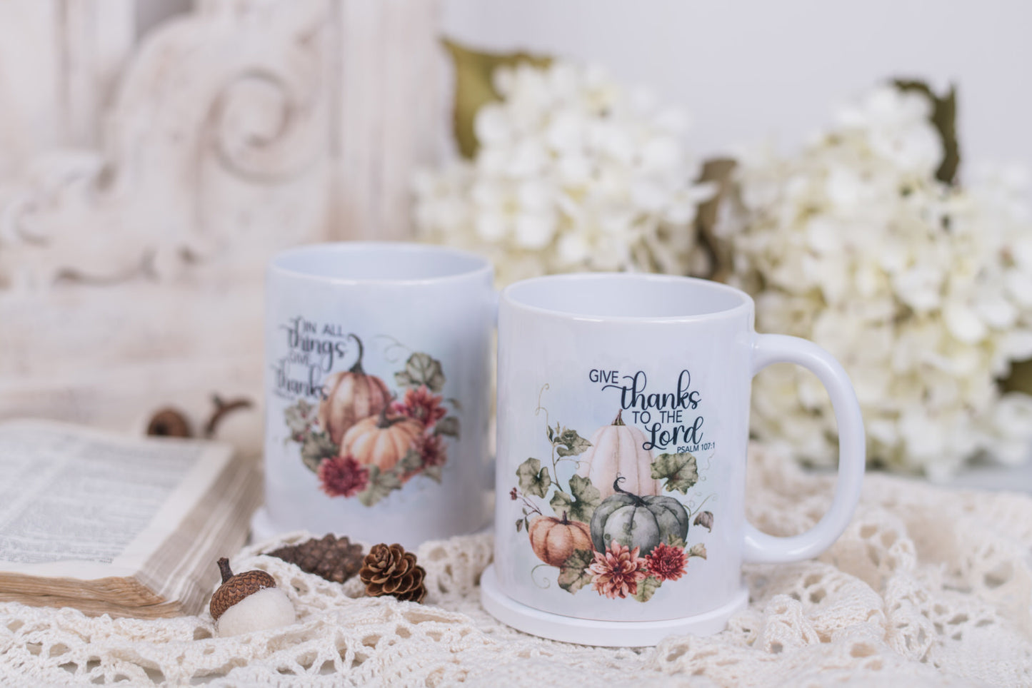Two mugs with autumn-themed designs and bible scripture on a decorative surface with flowers and an open book.
