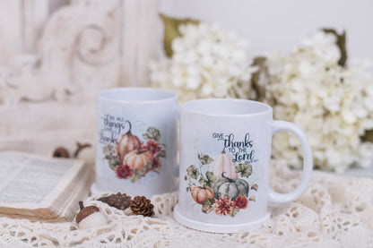 Two mugs with autumn-themed designs and bible scripture on a decorative surface with flowers and an open book.