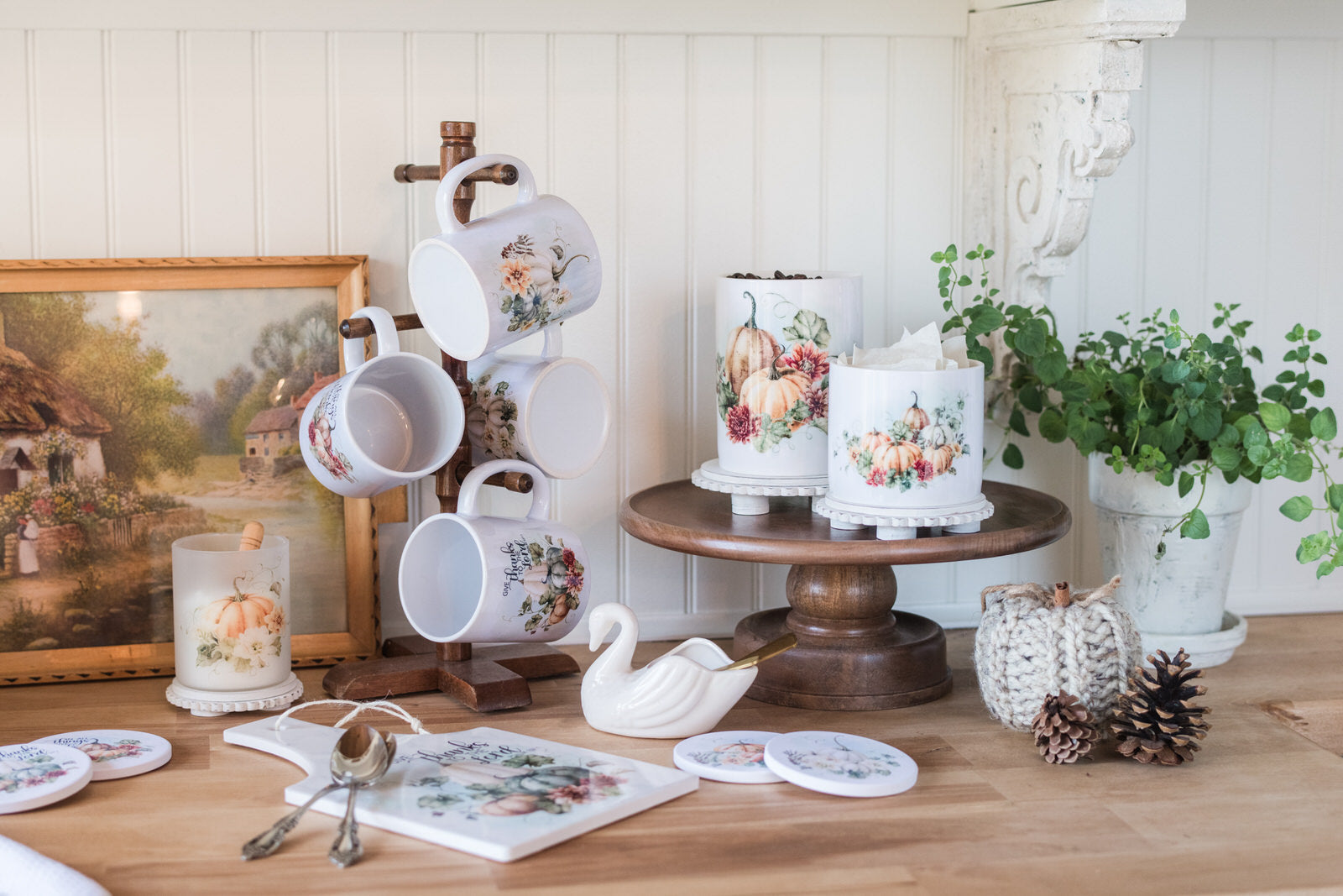 Fall kitchen items, including mugs, a cake stand with canisters and a painting on a wooden table.