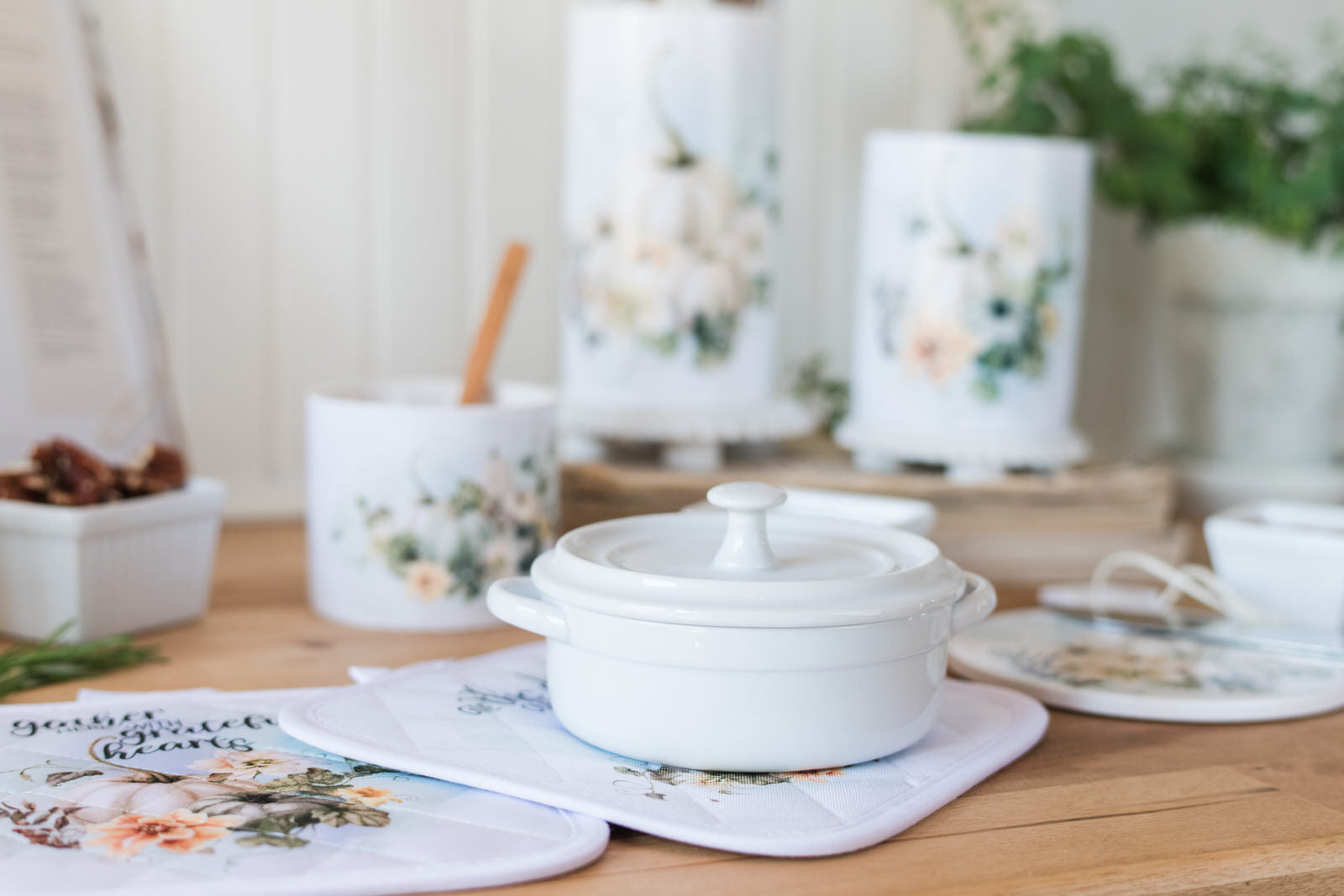 White ceramic pot sitting on fall pot holders, on a wooden table with pumpkins and a floral design and greenery in the background