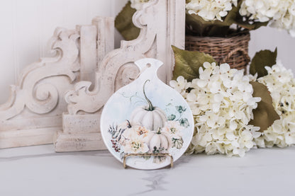 Pretty spoon rest with pumpkins and flowers on a counter with floral and wooden decor in the background.