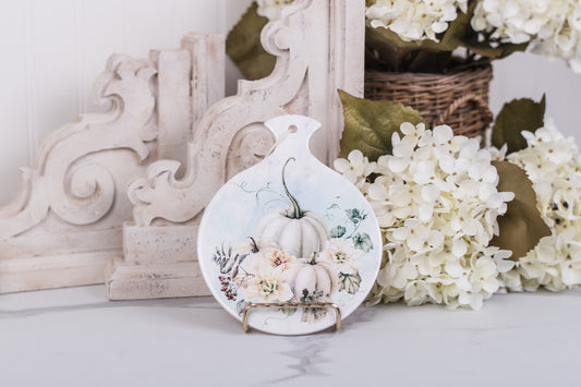 Pretty spoon rest with pumpkins and flowers on a counter with floral and wooden decor in the background.