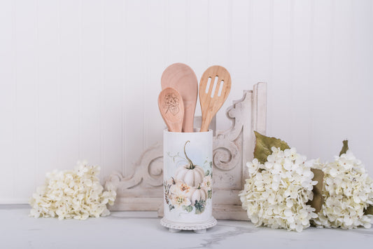Wooden utensils in a decorative container with pumpkins on a white surface with flowers.