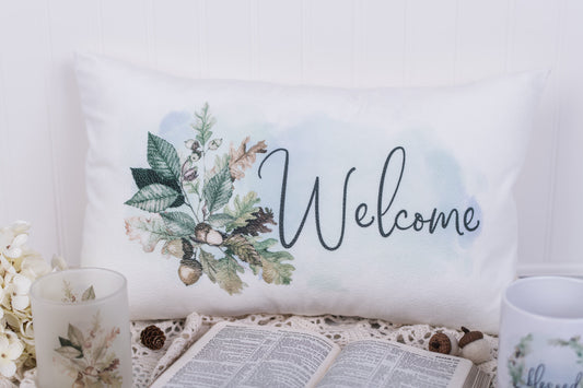 Decorative pillow with floral design and 'Welcome' text on a table with a candle and mug.