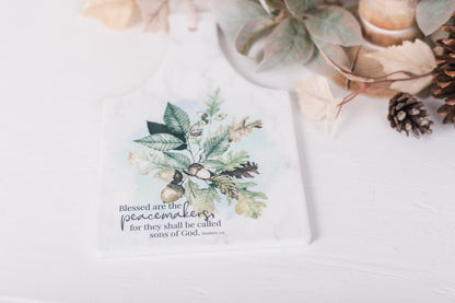 White ceramic trivet with scripture for Matthew 5:9 with fall leaves, and acorns styled on a countertop  
