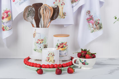 Kitchen utensils, mugs, and strawberries on a table with decorative towels in the background.