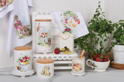 Decorative kitchen canisters with strawberry designs on a white surface with plants in the background.