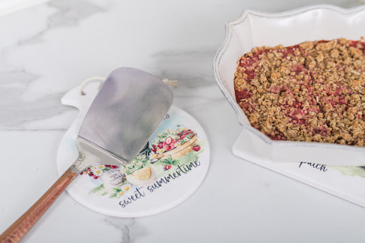 Baking dish with crumble dessert and spatula on a decorative mat with 'sweet summertime' text.
