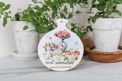 Watercolour strawberry spoon rest 
with floral design and 'sweet summertime' text on a surface with plants in the background.