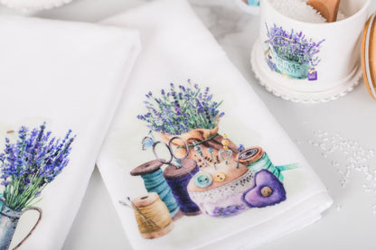 Decorative towel with a design of a pin cushion, lavender, and thread spools on a white surface.