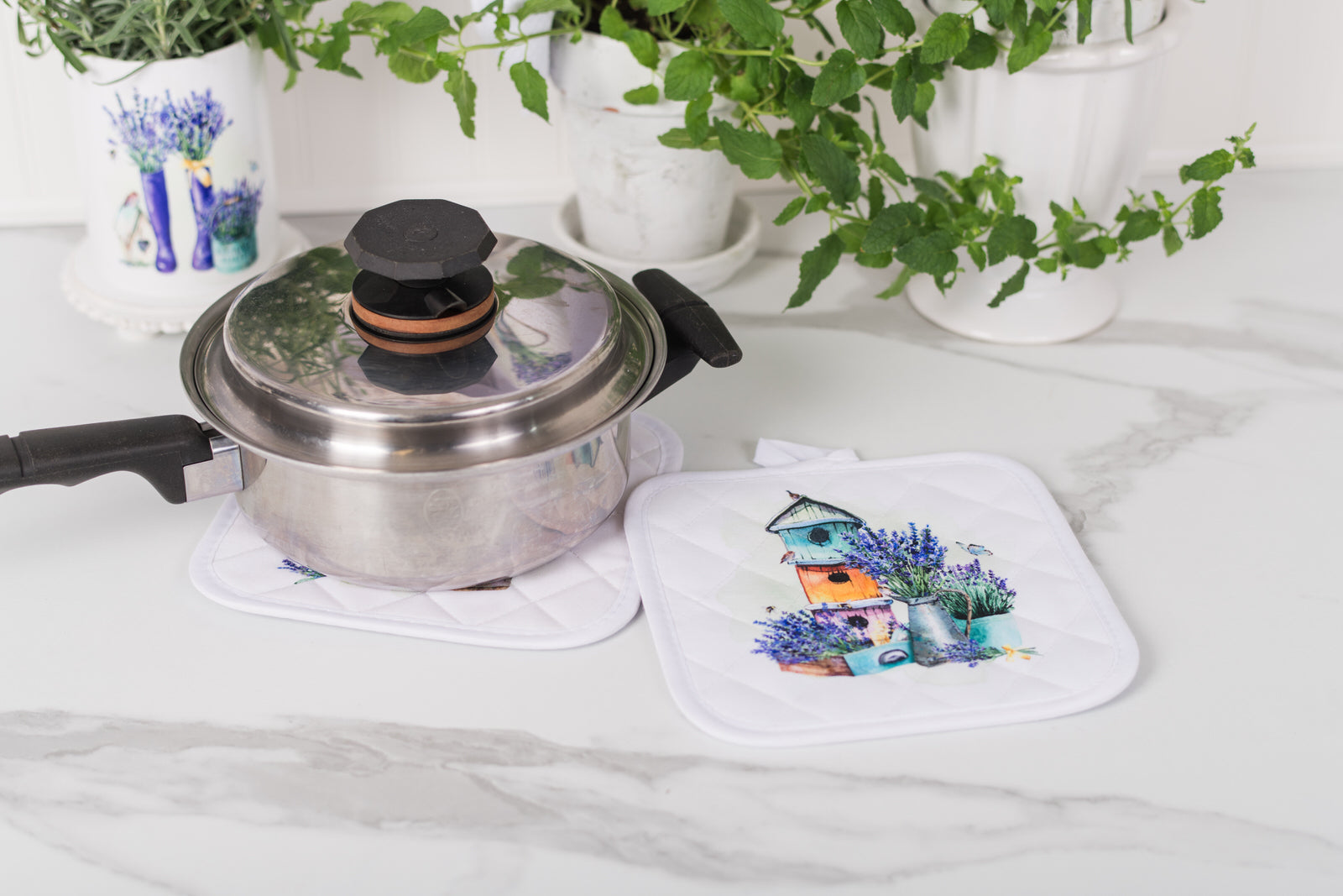 Stainless steel pot with a lid on a white surface, next to a matching potholder with a garden design.