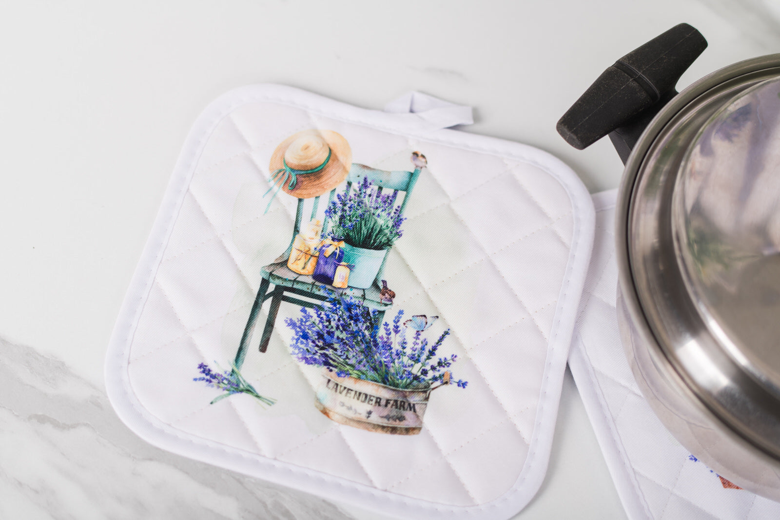 Stainless steel pot with a decorative potholder featuring a chair and lavender design on a white surface.