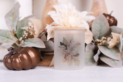 A frosted glass candle jar with a bamboo lid, featuring a leaf pattern with autumn colours.