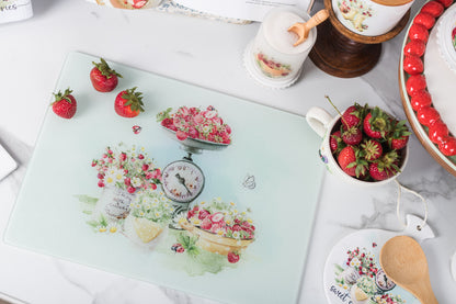 Decorative glass cutting board with fruit design on a table with tea set and strawberries.