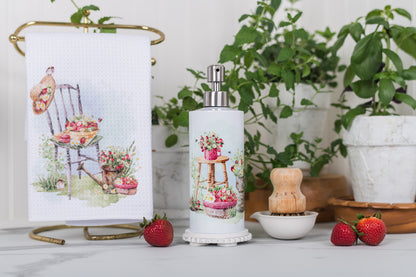 Decorative soap dispenser with a printed label featuring a chair and flowers, surrounded by plants and strawberries on a light surface.